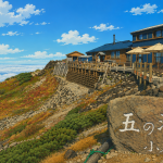 Rock in foreground engraved with Japanese text ‘五の池小屋’; blue mountain cafe building with deck and umbrellas on a rocky hillside above clouds