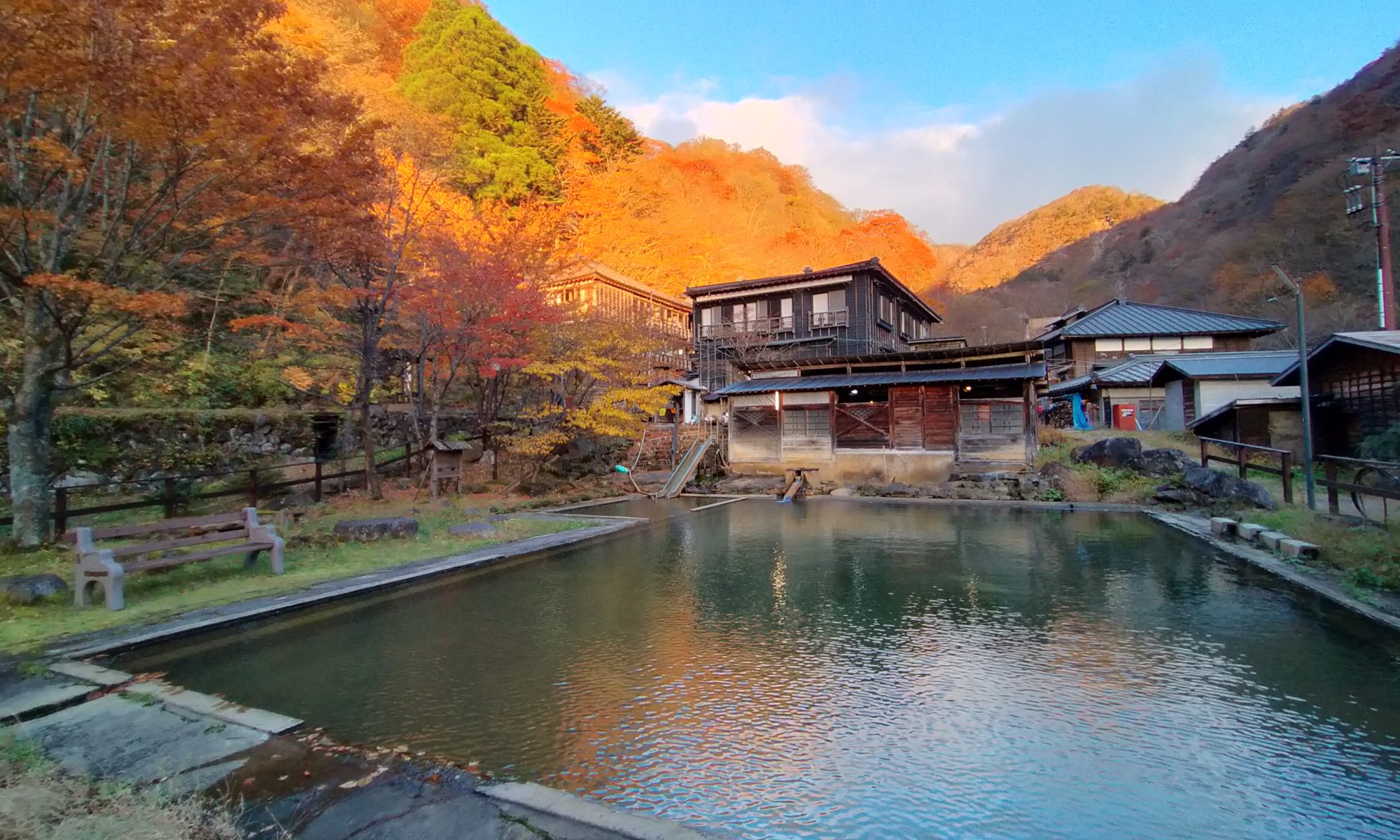 Autumn scene of a tranquil pond with traditional wooden buildings, orange trees, and sunlit hills in the background.