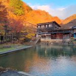 Autumn scene of a tranquil pond with traditional wooden buildings, orange trees, and sunlit hills in the background.