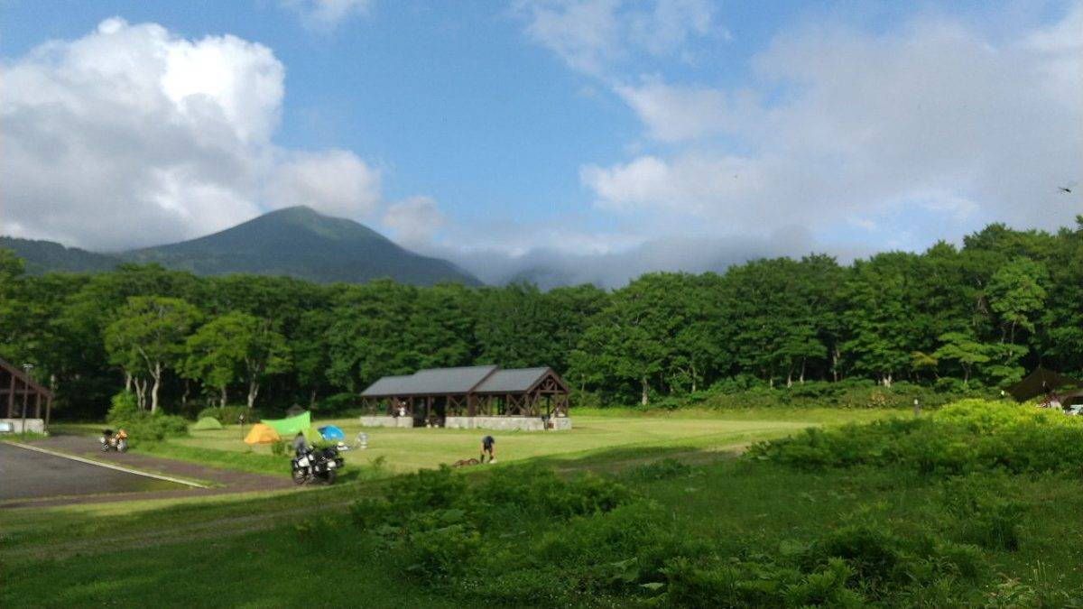 Campsite on a grassy field with colorful tents, a wooden shelter, and forested hills under a blue sky with clouds