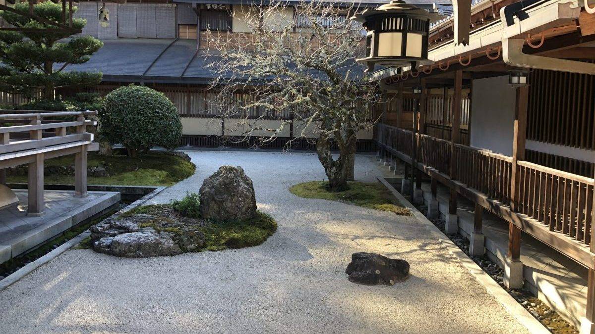 Traditional Japanese temple courtyard with wooden walkways, a hanging lantern, and a raked gravel ground bordered by rocks and neatly trimmed greenery.